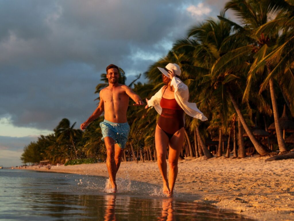 Couple running hand-in-hand along a palm-lined tropical beach at sunset, splashing through shallow water.